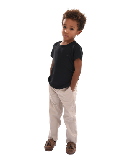 A boy with curly hair looking at the camera. He is wearing clothing from Moustache Kids. A black t-shirt, beige pants and coffee brown moccasin dress shoes. 