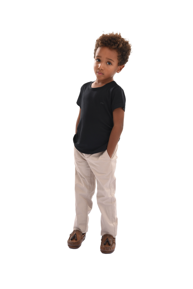 A boy with curly hair looking at the camera. He is wearing clothing from Moustache Kids. A black t-shirt, beige pants and coffee brown moccasin dress shoes. 