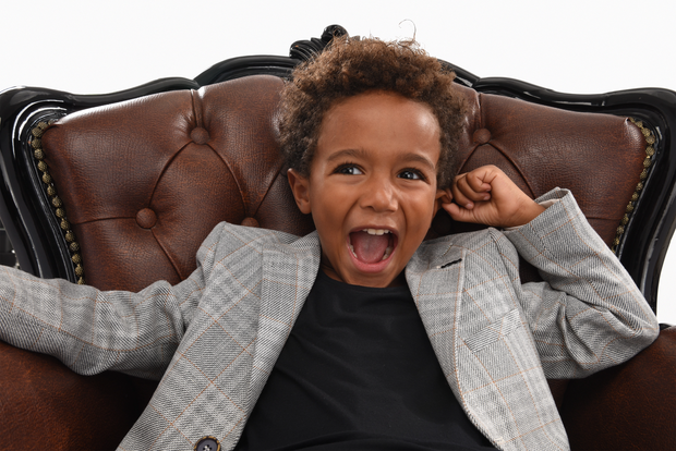 A Moustache Kids model with curly hair wearing a plaid blazer and black t shirt laughing and looking off camera.